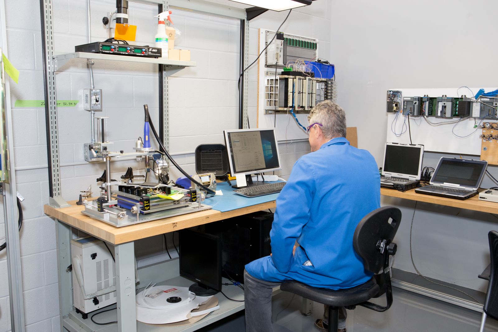 AST technician at an electronics repair bench with a circuit board rework station, test computer, and PLC racks mounted on the wall